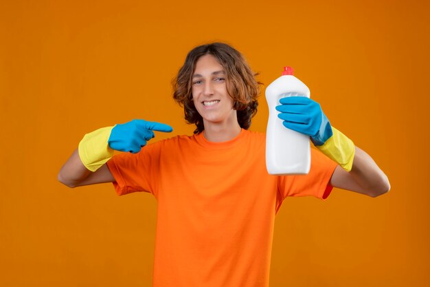 Giovane uomo in maglietta arancione che indossa guanti di gomma tenendo una bottiglia di prodotti per la pulizia puntando con il dito ad esso sorridendo allegramente in piedi su sfondo giallo