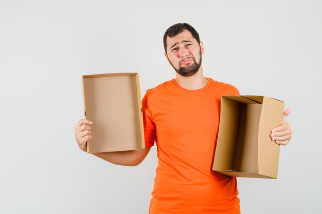 Giovane uomo in maglietta arancione che tiene in mano una scatola di cartone vuota e guarda abbattuto, vista frontale.