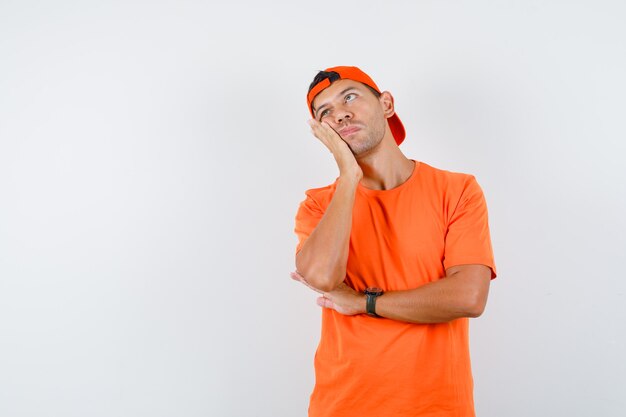 Giovane uomo in maglietta arancione e berretto appoggiato la guancia sul palmo sollevato e guardando pensieroso
