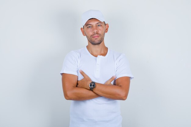 Giovane uomo in maglietta bianca, cappello in piedi con le braccia incrociate, vista frontale.