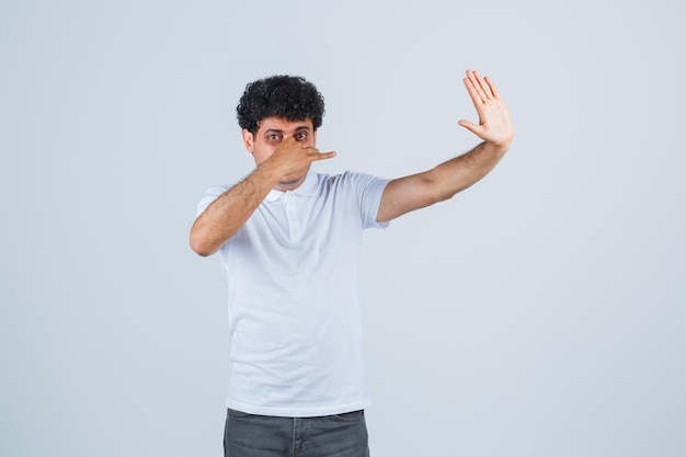 Giovane uomo in maglietta bianca e jeans che pizzica il naso a causa del cattivo odore, mostra il segnale di stop e sembra tormentato, vista frontale.