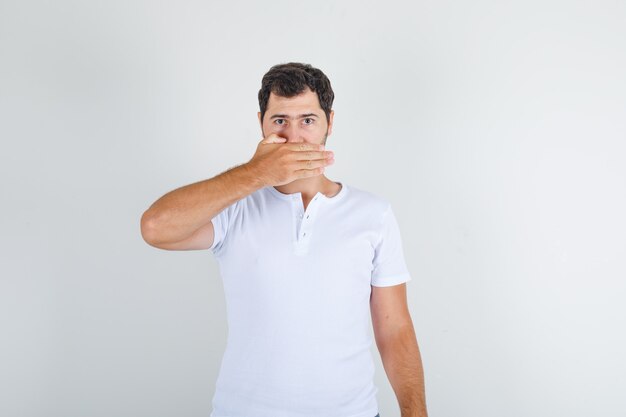 Giovane uomo in maglietta bianca tenendo la mano sulla bocca e guardando la calma