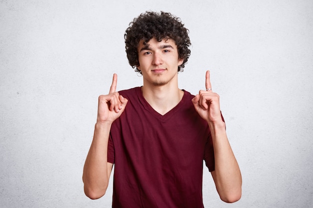 giovane uomo in maglietta casual marrone, che punta il dito indice, mostrando qualcosa, isolato sul muro bianco.