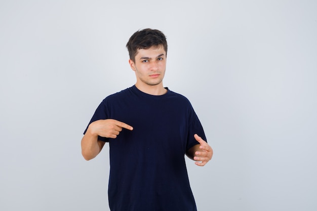 Giovane uomo in maglietta nera che punta al palmo della mano allargato e guardando serio, vista frontale.