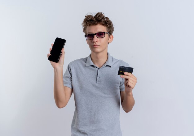 Giovane uomo in maglietta polo grigia che mostra smartphone tenendo la carta di credito con la faccia seria in piedi sopra il muro bianco