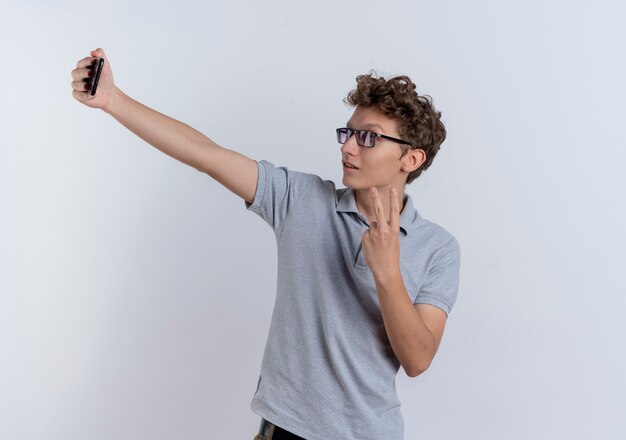 Giovane uomo in maglietta polo grigia guardando il suo schermo dello smartphone facendo selfie che mostra v-sign in piedi sopra il muro bianco