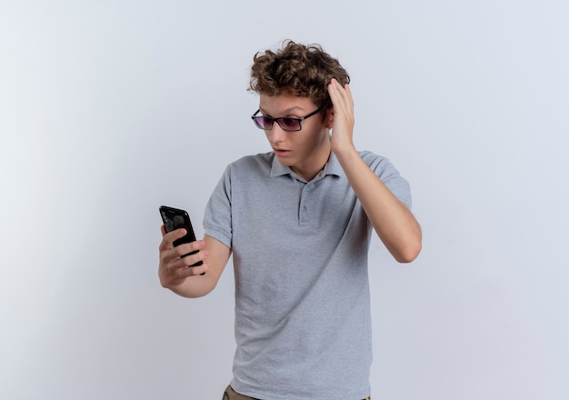 Giovane uomo in maglietta polo grigia guardando lo schermo dello smartphone essendo confuso in piedi sopra il muro bianco