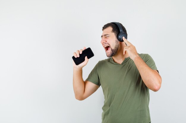 Giovane uomo in maglietta verde che canta nel cellulare come microfono e guardando comico