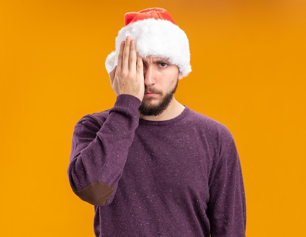 Giovane uomo in maglione viola e cappello da Babbo Natale che copre un occhio con la mano in piedi su sfondo arancione