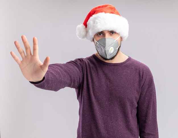 Giovane uomo in maglione viola e cappello da Babbo Natale che indossa la maschera protettiva per il viso che guarda l'obbiettivo con faccia seria che fa gesto di arresto con la mano in piedi su sfondo bianco