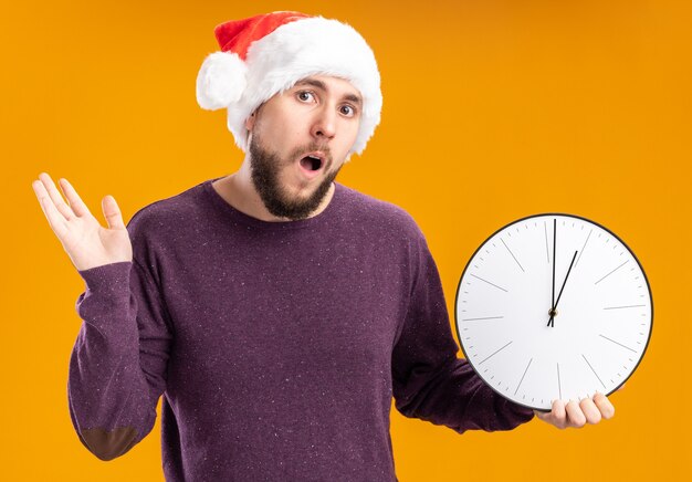 Giovane uomo in maglione viola e cappello da Babbo Natale che tiene orologio da parete confuso e molto ansioso scrollare le spalle in piedi sopra la parete arancione