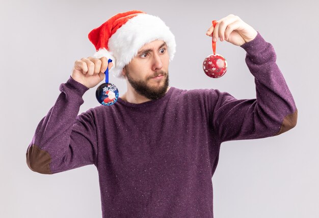 Giovane uomo in maglione viola e cappello da Babbo Natale indossando occhiali divertenti che tengono le palle di Natale guardandoli confusi cercando di fare la scelta in piedi sul muro bianco