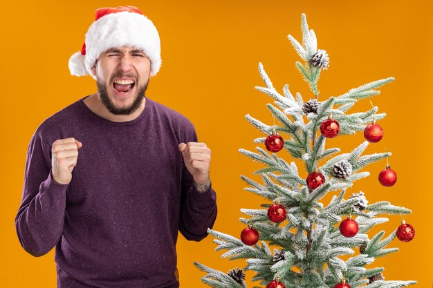 Giovane uomo in maglione viola e cappello da Babbo Natale stringendo i pugni gridando con espressione aggressiva in piedi accanto all'albero di Natale su sfondo arancione