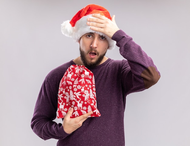 Giovane uomo in maglione viola e cappello da Babbo Natale tenendo la borsa rossa con doni cercando confuso con la mano sulla sua testa, dimenticato in piedi su sfondo bianco