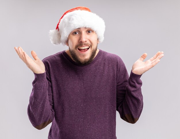 Giovane uomo in maglione viola e cappello santa guardando la telecamera sorridendo confuso diffondendo le braccia ai lati in piedi su sfondo bianco