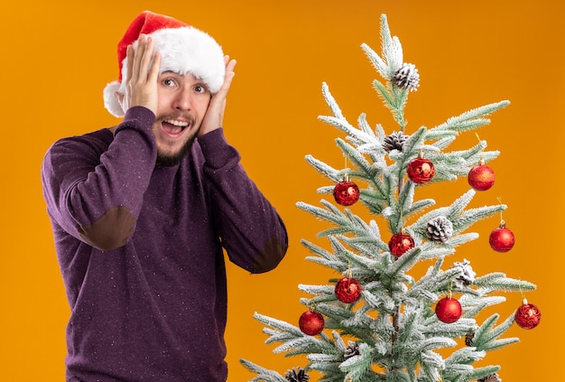 Giovane uomo in maglione viola e cappello santa guardando stupito e sorpreso in piedi accanto all'albero di Natale su sfondo arancione