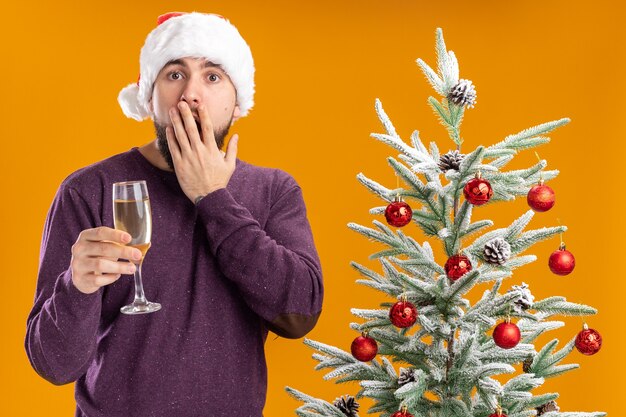 Giovane uomo in maglione viola e cappello santa tenendo un bicchiere di champagne stupito e sorpreso in piedi accanto all'albero di Natale sopra la parete arancione