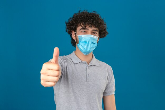 Giovane uomo in maschera protettiva medica che guarda l'obbiettivo con la faccia felice che mostra il pollice in su su sfondo blu isolato