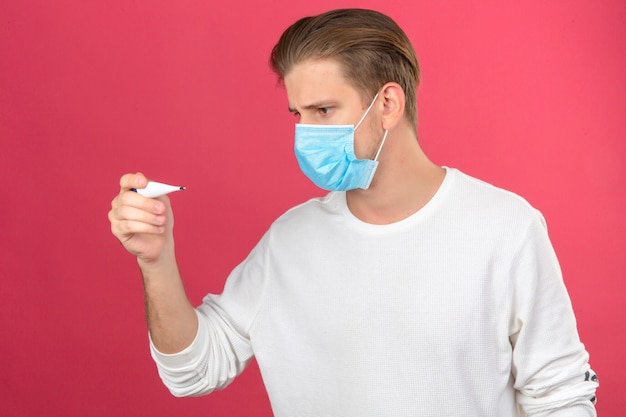 Giovane uomo in maschera protettiva medica guardando il termometro digitale in preda al panico scioccato e sorpreso su sfondo rosa isolato