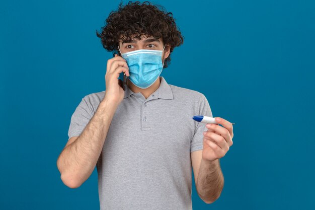 Giovane uomo in maschera protettiva medica parlando al telefono cellulare tenendo il termometro digitale in altra mano cercando nervoso su sfondo blu isolato