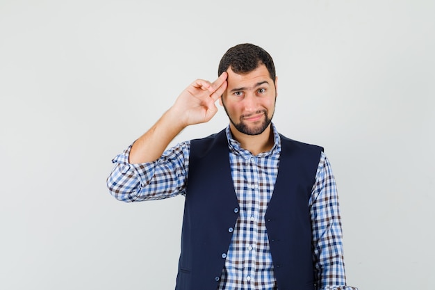 Giovane uomo in posa con le dita puntate alla testa in camicia, gilet e guardando fiducioso.