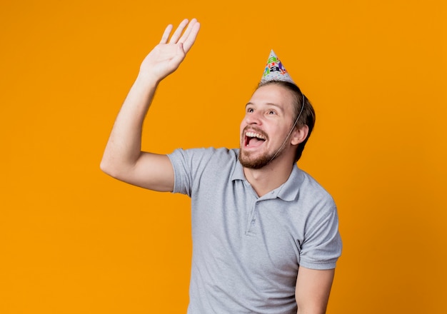 Giovane uomo in protezione di vacanza che osserva da parte agitando con la mano sorridendo allegramente festa di compleanno concetto in piedi sopra la parete arancione