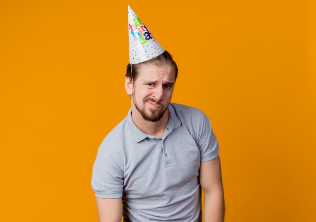 Giovane uomo in protezione di vacanza che sembra scontento concetto di festa di compleanno in piedi sopra la parete arancione