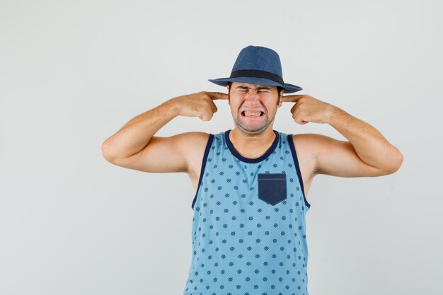 Giovane uomo in singoletto blu, cappello tappando le orecchie con le dita e guardando infastidito