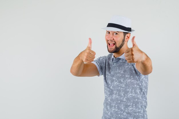 Giovane uomo in t-shirt, cappello che mostra il doppio pollice in alto e sembra felice, vista frontale.