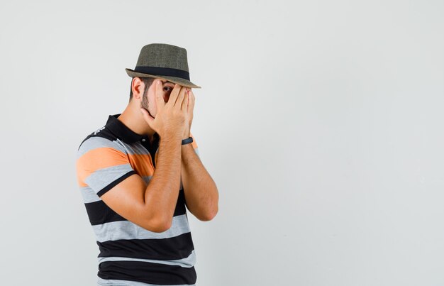 Giovane uomo in t-shirt, cappello guardando attraverso le dita e guardando spaventato, vista frontale.