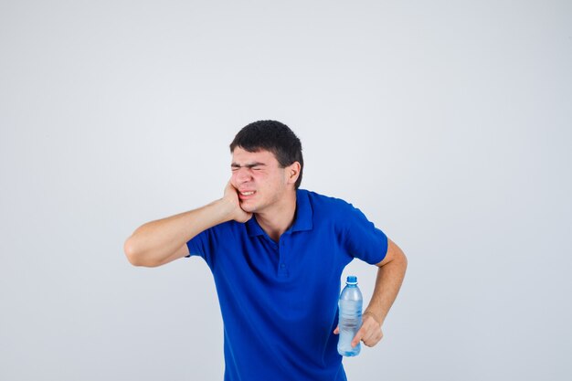 Giovane uomo in t-shirt che soffre di mal di denti, che tiene la bottiglia di plastica e che sembra doloroso, vista frontale.