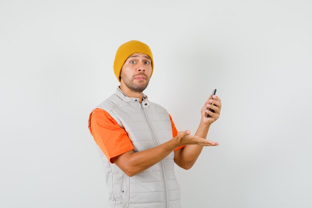 Giovane uomo in t-shirt, giacca, cappello che tiene il telefono cellulare e che sembra confuso