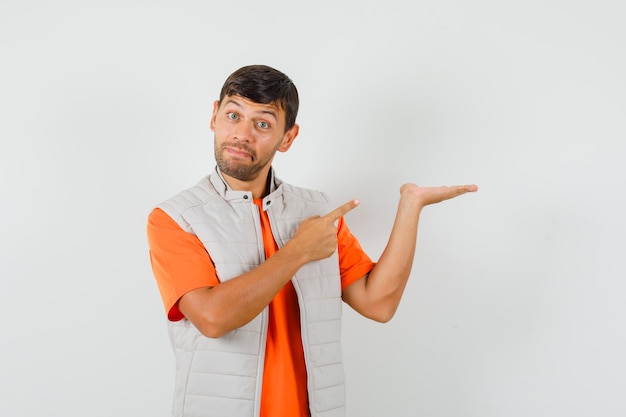 Giovane uomo in t-shirt, giacca che punta da parte, fingendo di tenere qualcosa d'altra parte, vista frontale.