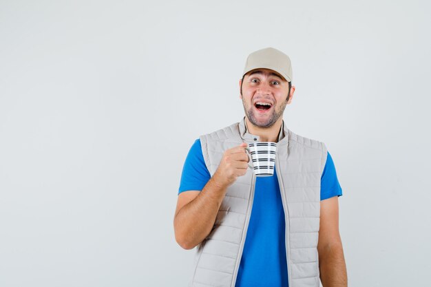 Giovane uomo in t-shirt, giacca che tiene tazza di bevanda e che sembra felice, vista frontale.