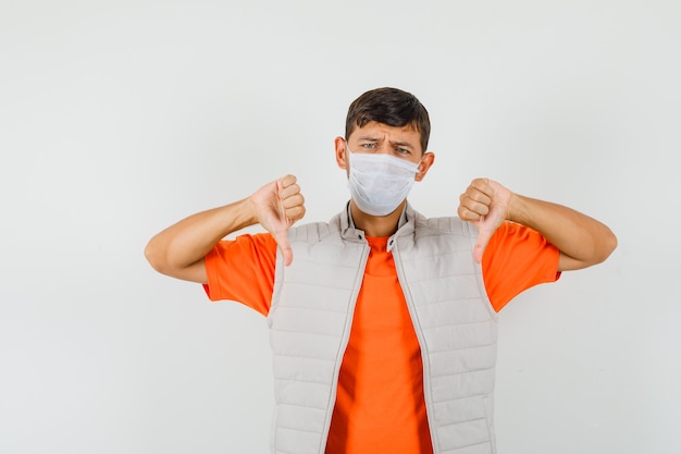 Giovane uomo in t-shirt, giacca, maschera che mostra i doppi pollici verso il basso e sembra deluso, vista frontale.