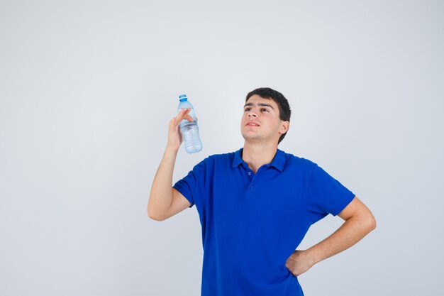 Giovane uomo in t-shirt mantenendo la bottiglia di plastica, tenendo la mano sulla vita e guardando fiducioso, vista frontale.