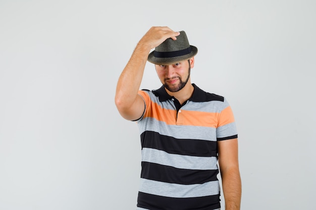 Giovane uomo in t-shirt tenendo la mano sul suo cappello e sorridente, vista frontale.