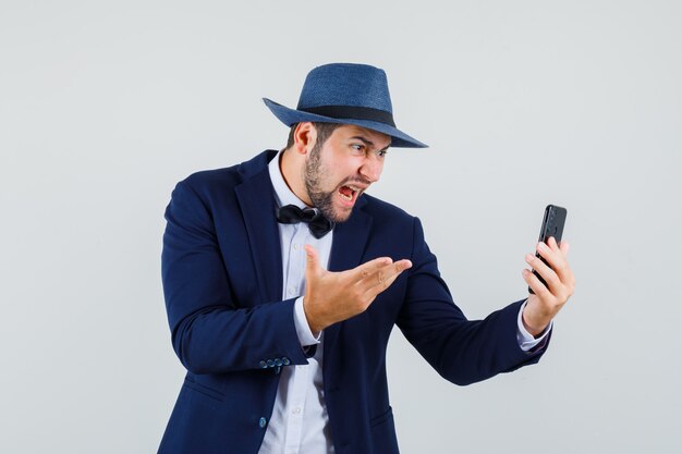 Giovane uomo in tuta, cappello che grida mentre si effettua una videochiamata e sembra arrabbiato, vista frontale.