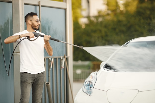 Giovane uomo in vestiti di moda che lava l'auto alla stazione di autolavaggio utilizzando una macchina per l'acqua ad alta pressione. Uomo che indossa una maglietta bianca e jeans