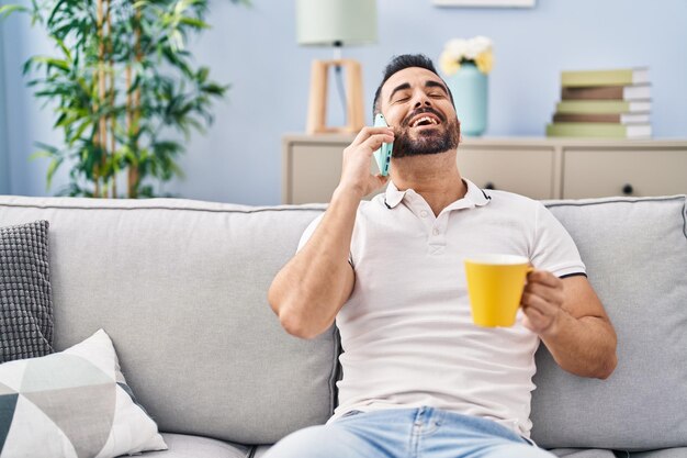 Giovane uomo ispanico che beve caffè e parla sullo smartphone a casa