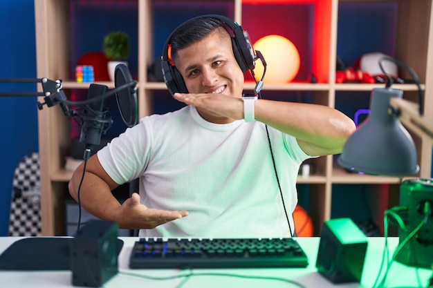 Giovane uomo ispanico che gioca ai videogiochi gesticolando con le mani che mostrano il simbolo di misura del segno di grandi e grandi dimensioni sorridendo guardando il concetto di misurazione della fotocamera