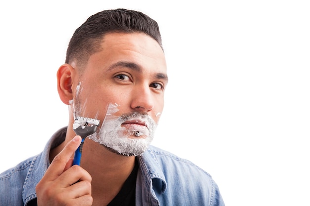 Giovane uomo ispanico che guarda la telecamera come se fosse uno specchio e usa un rasoio per radersi la barba su sfondo bianco