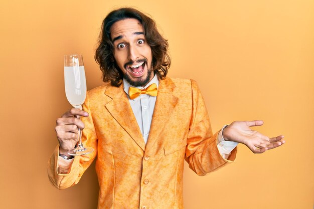 Giovane uomo ispanico che indossa uno stile elegante da nerd bevendo un bicchiere di champagne frizzante che celebra il successo con un sorriso felice e l'espressione del vincitore con la mano alzata