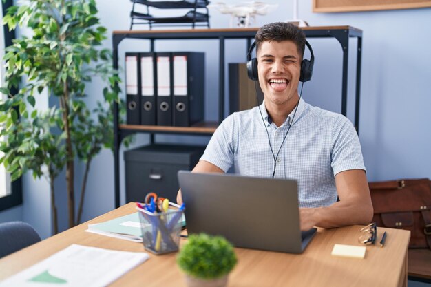 Giovane uomo ispanico che lavora in ufficio indossando le cuffie con la lingua fuori felice con il concetto di emozione di espressione divertente