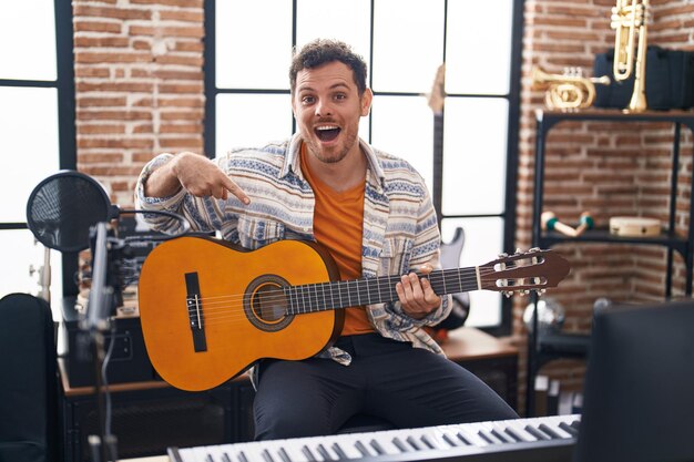 Giovane uomo ispanico che suona la chitarra classica in studio musicale sorridendo felice indicando con mano e dito