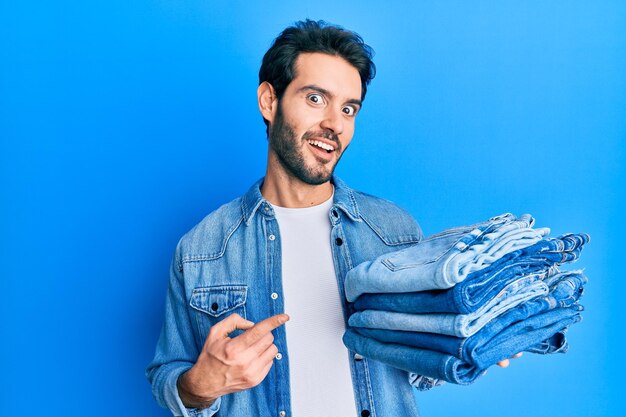 Giovane uomo ispanico che tiene pila di jeans piegati sorridendo felice indicando con mano e dito