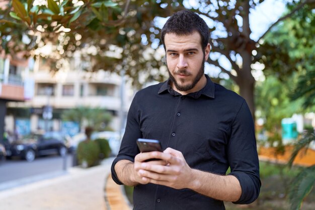 Giovane uomo ispanico sorridente fiducioso utilizzando lo smartphone al parco