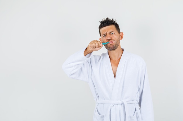 Giovane uomo lavarsi i denti con le sopracciglia aggrottate la fronte in vista frontale accappatoio bianco.