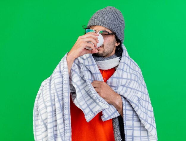 Giovane uomo malato caucasico con gli occhiali cappello invernale e sciarpa avvolti in plaid afferrando plaid guardando all'interno tazza bere tè isolato sul muro verde