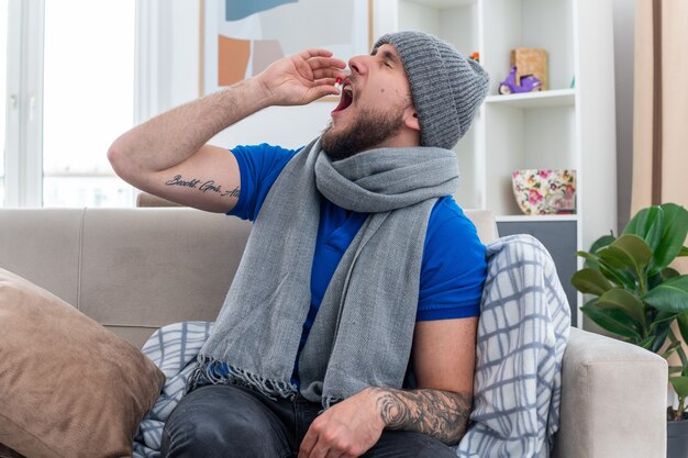 Giovane uomo malato che indossa sciarpa e cappello invernale seduto sul divano in soggiorno prendendo capsula medica con gli occhi chiusi